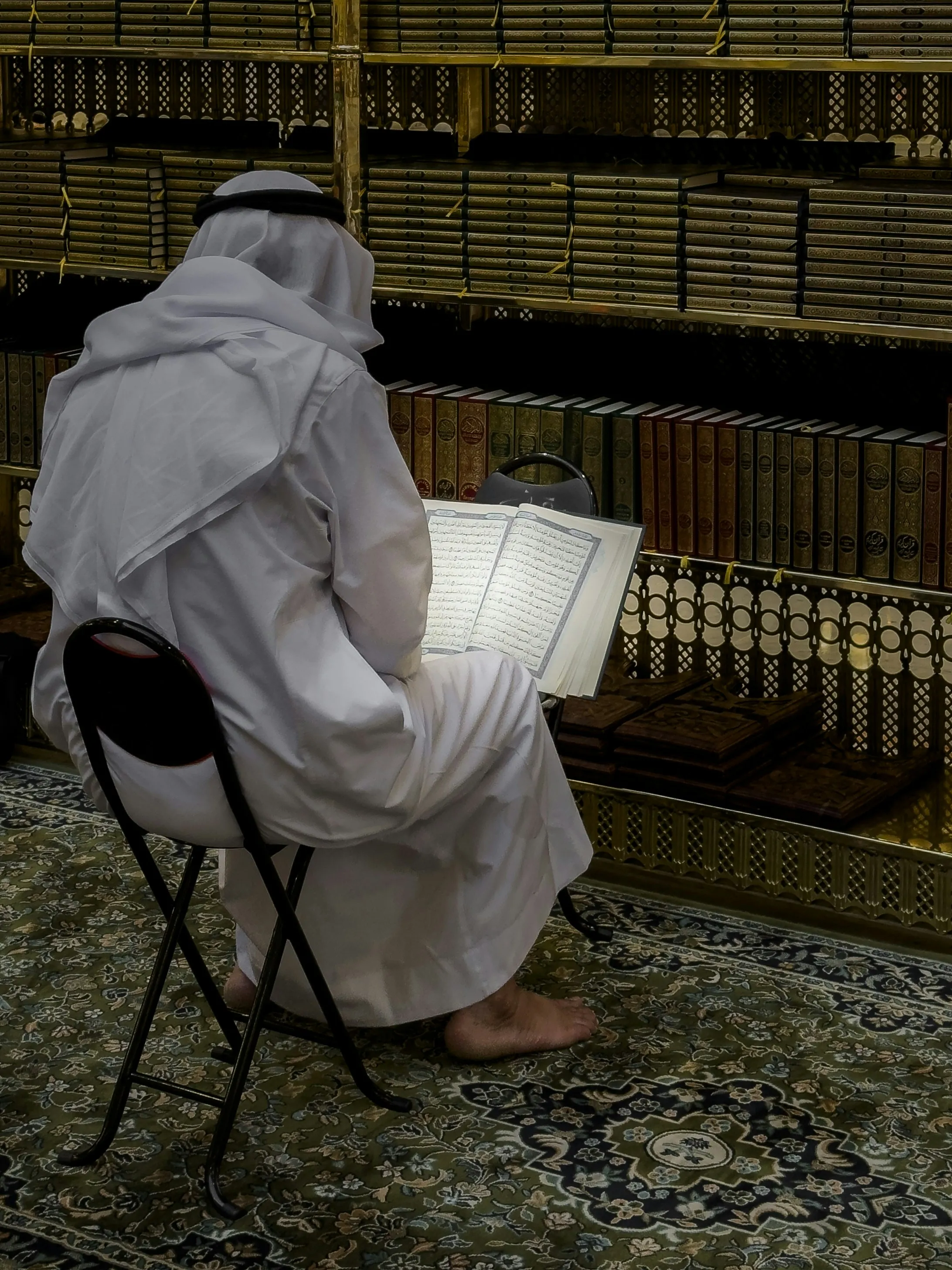 Een man in wit gewaad zit op een klapstoeltje in een bibliotheek vol boekenkasten en leest in een opengeslagen mushaf