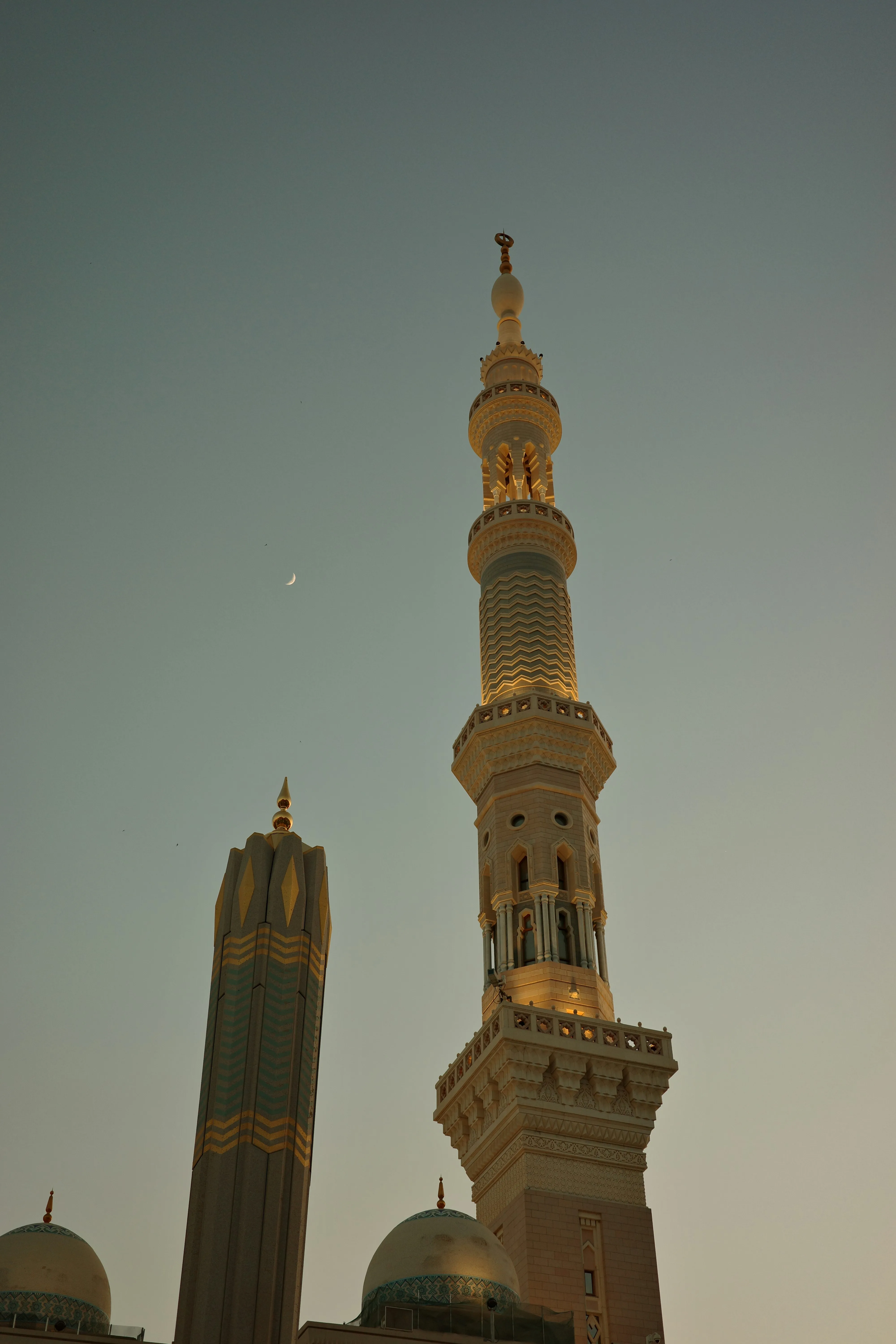 Verlichte minaret bij schemering, met een smalle maansikkel aan de lucht en koepels van de moskee eronder